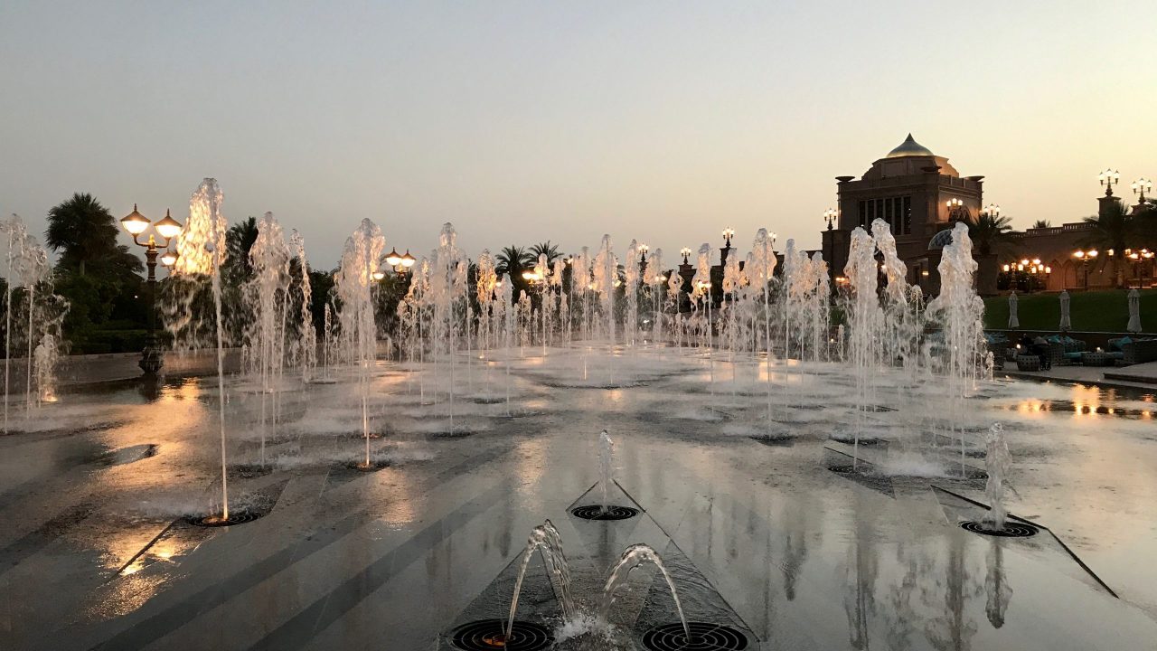Wasserspiele beim Sonnenuntergang. Im Hintergrund sind die vielen Lampen und ein Teil der Außenfassade vom Luxushotel Emirates Palace in Abu Dhabi bereits erleuchtet. Lichter spiegeln sich im Wasser.