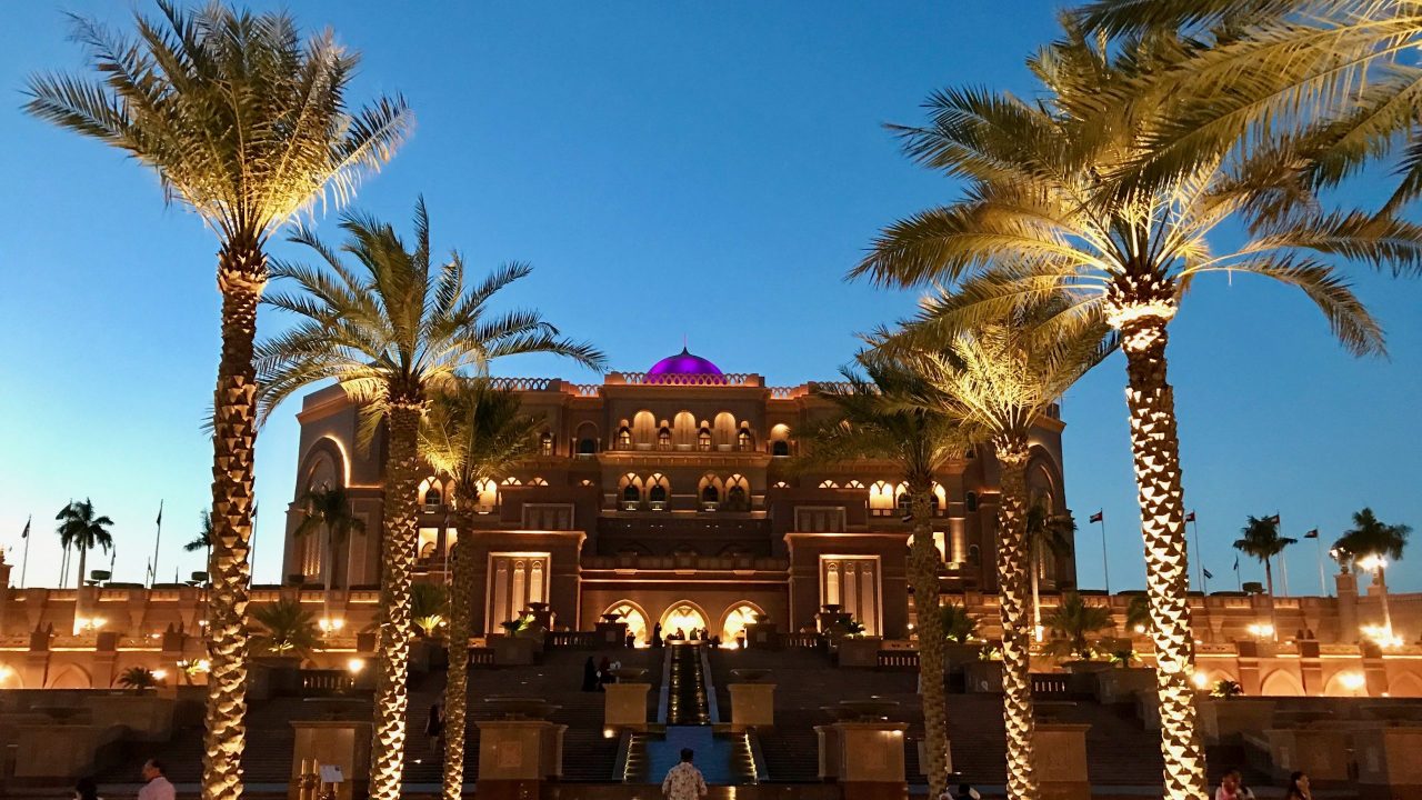 Einmalige Opulenz und ein Palast wie aus einem Märchen, das Luxushotel Emirates Palace in Abu Dhabi. Außenfassade überdimensional groß, von Palmen gesäumte Stufen zum Eingang. Blauer Nachthimmel wird von einem Lichtermeer zum Leuchten gebracht.