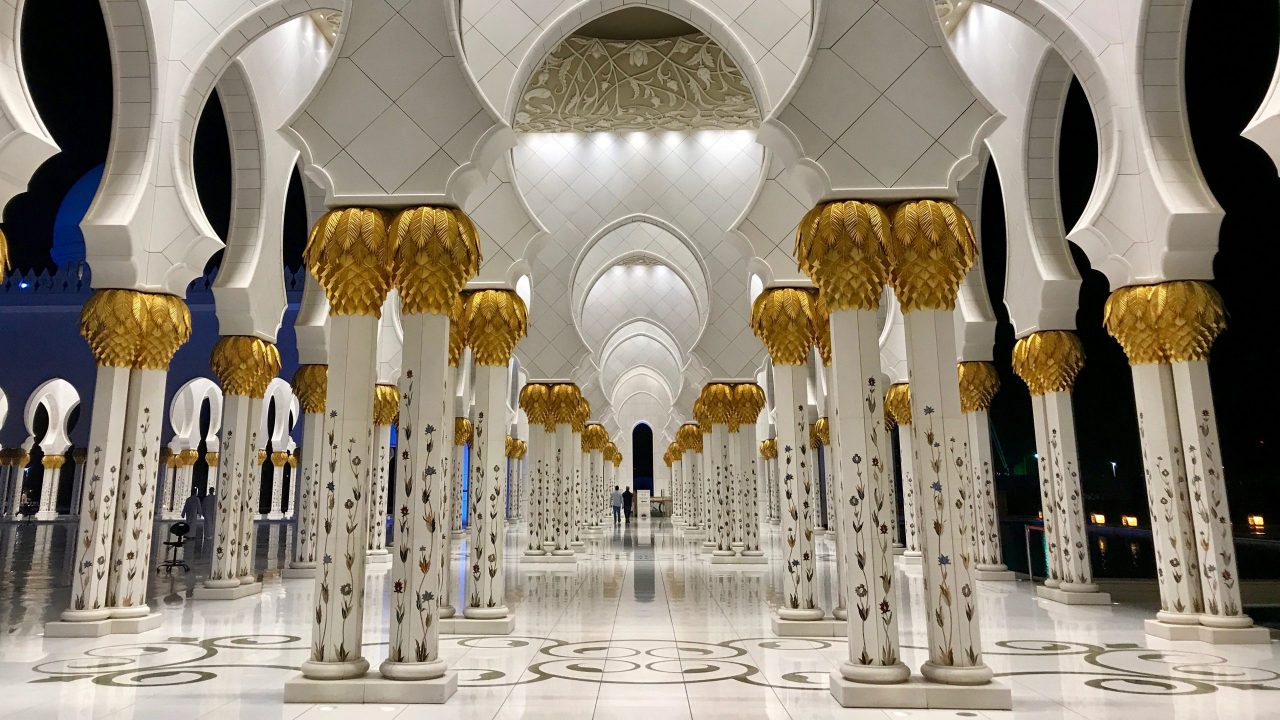 Scheich-Zayid-Moschee Abu Dhabi bei Nacht. Innen leuchtet die Mosche in hellem Kunstlicht. Das Gold der weißen Säulen glänzt vor dem dunklen Himmel. Der weiße Marmorboden strahlt und glänzt.