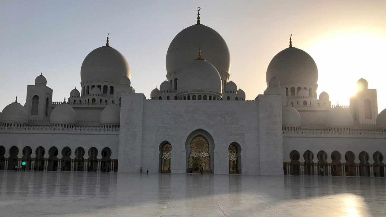 Scheich-Zayid-Moschee in Abu Dhabi beim Sonnenuntergang. Die weiße Außenfassade ist leicht im Schatten, wunderschöne Schattenspiegelungen sendet die untergehende goldgelbe Sonne auf dem blitzblanken Marmorboden.