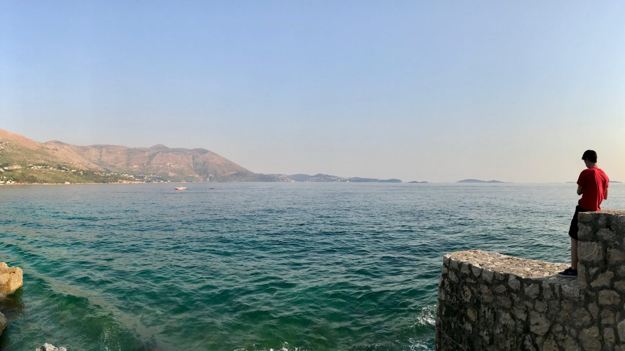 Türkisblaues Meer, im Hintergrund kahle Berge und grüne Natur. Ein Kind blickt auf das offene Meer hinaus