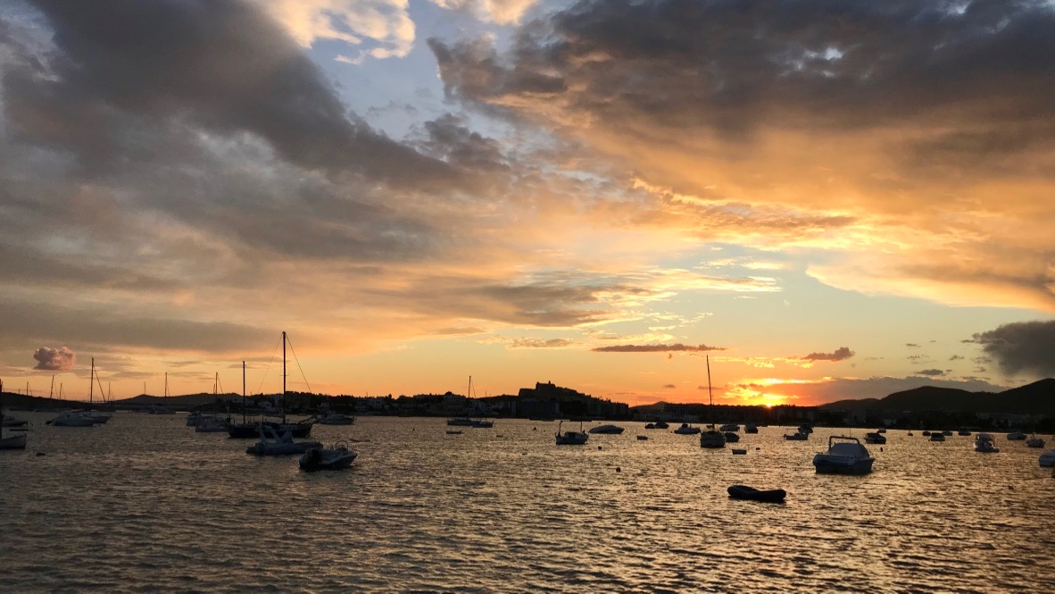 Ibiza Hafen im Sonnenuntergang. Die Altstadt leuchtet und dahinter wirft die orangegelbe Sonne ihre Licher über den Himmel.