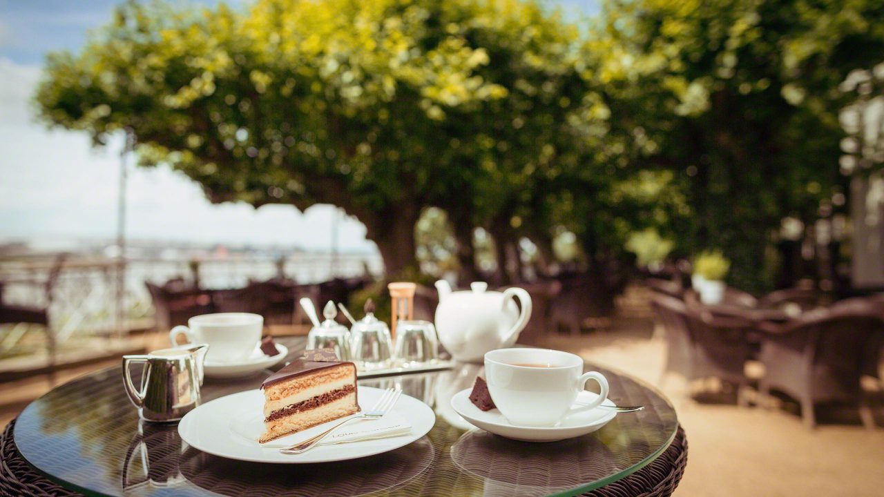 Lindenterrasse des Hotel Louis C. Jacob Hamburg. Der Tisch ist eingedeckt mit Tee und Champagnertorte mit Blick auf die grünen Bäume und die Elbe samt Hafen.